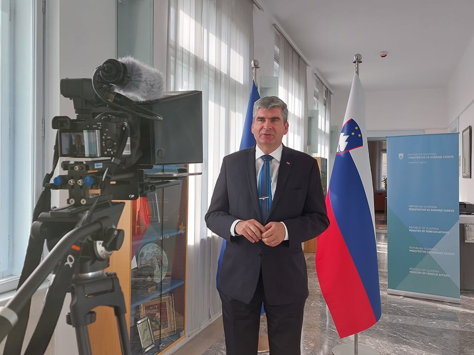 state secretary stands infront of flag, looks into camera