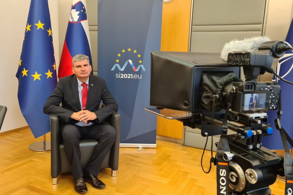 Državni sekretar Stanislav Raščan sedi, gleda v kamero,  ozadju zastave