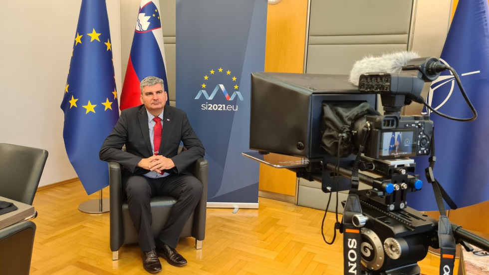 Državni sekretar Stanislav Raščan sedi, gleda v kamero,  ozadju zastave