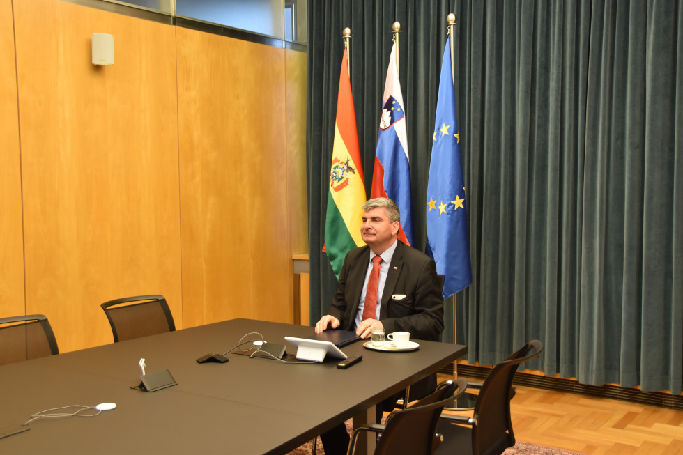 državni sekretar sedi za mizo, v ozadju zastave Bolivije, Slovenije in Evropske unije