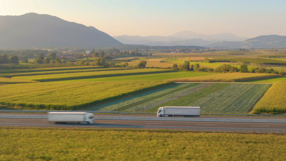 Posnetek iz zraka dveh tovornjakov, ki prevažata tovor po slikoviti avtocesti, ki prečka živahno zeleno slovensko pokrajino.