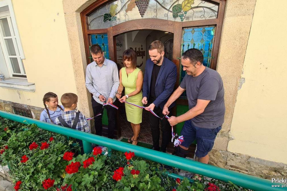 Gostje stojijo pred vhodnimi vrati v trgovino in režejo trak, na levi dva otroka, ki pomagata trak pridržati.