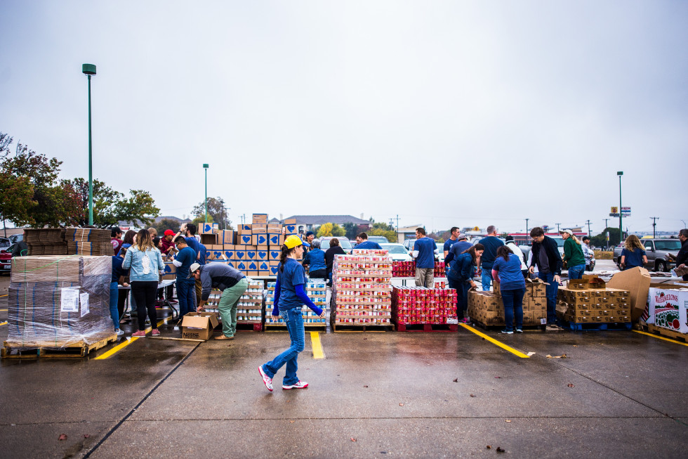 Na fotografiji je parkirišče, kjer več ljudi sortira in organizira škatle z različnimi dobrinami za nadaljnjo distribucijo. 