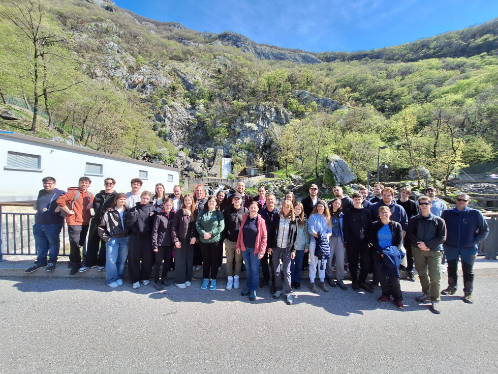 Mladi stojijo eden zraven drugega zunaj na soncu. Za njimi rahla vzpetina in drevesa.