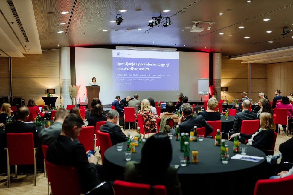 Udeleženci sedijo za okroglimi mizami. Na odru veliko platno, na katerem se odvija predstavitev.