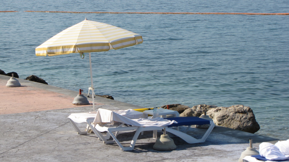 Slika je okrasna. Na njej je betonirana plaža s senčnikom in ležalnikoma ter pogledom na morje