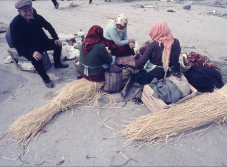 Diapozitiv Bojana Adamiča z upodobitvijo treh starejših žensk z rutami, ki sedijo na senu. Ob njih je na polomljenem lesenem zaboju starejši moški z brki in kapo.