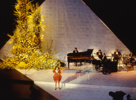 Diapozitiv Bojana Adamiča z upodobitvijo koncertnega nastopa. V ospredju je pevka v krdeči obleki, v ozadju glasbeniki za klavirjem, kitaro, bobni.
