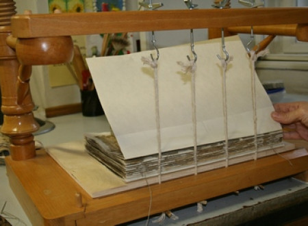 Šivanje knjižnega bloka rokopisne knjige, ki je vpeta v leseno stojalo.