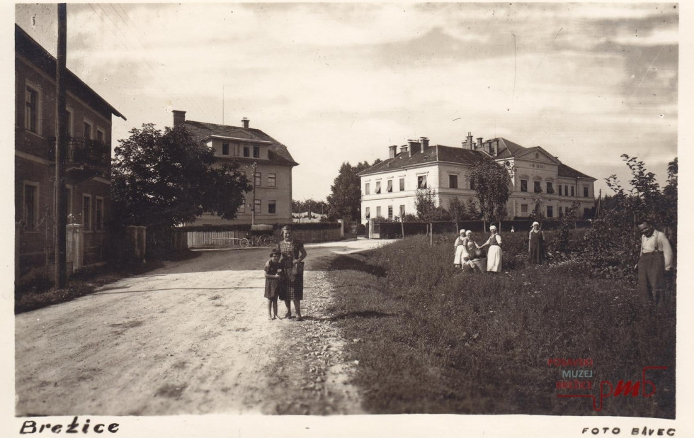 Črno-bela fotografija brežiške bolnišnice  z makadamsko potjo, ki vodi do nje, in prebivalci okoliških hiš.