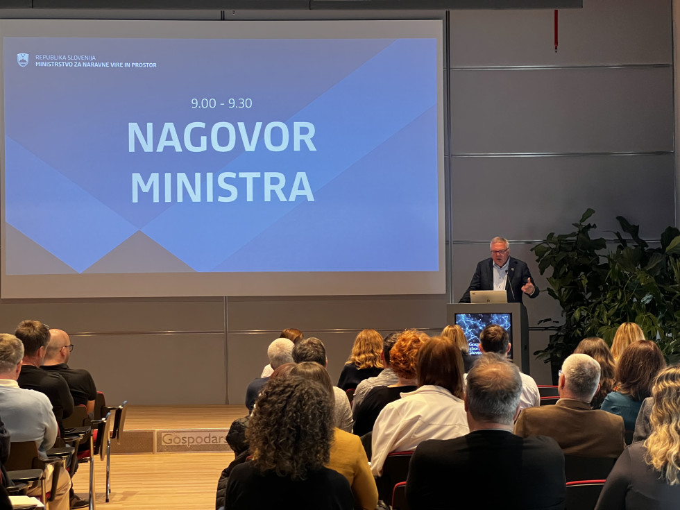 Moški za govorniškim pultom nagovarja udeležence v dvorani