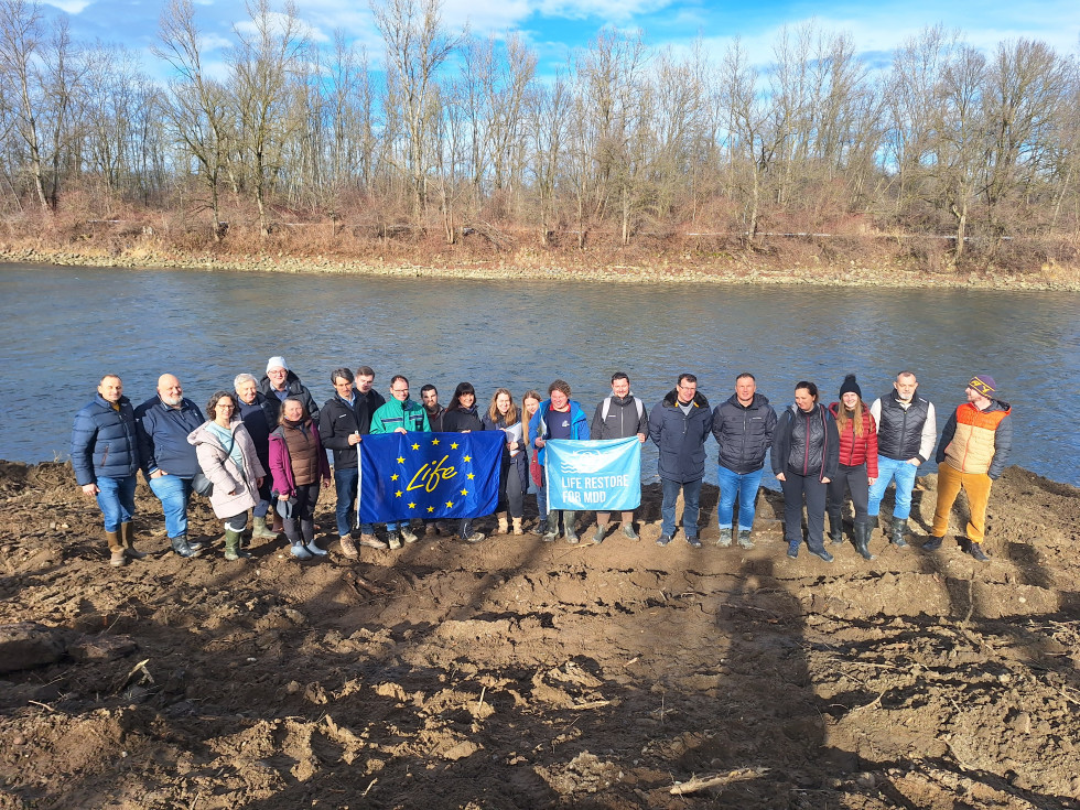 Udeleženci posveta pozirajo v vrsti pred vodotokom. V rokah držijo zastavo Life in Life Restore.