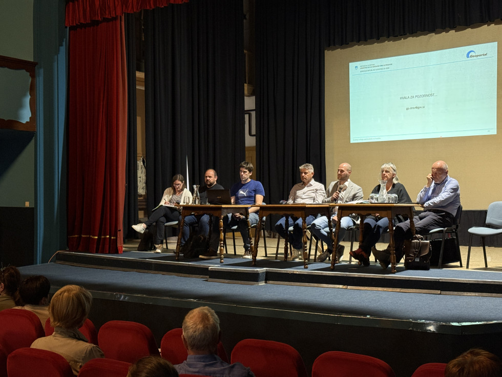 Panelna razprava na odru; govorniki sedijo za mizo, občinstvo spremlja dogodek v dvorani.