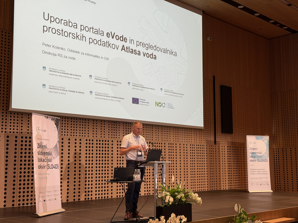 Oseba za govorniškim pultom nagovarja udeležence konference, v ozadju pa je projekcijsko platno s predstavitvijo.