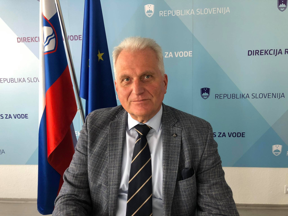 Moški sedi pred slovensko in evropsko zastavo, v ozadju stena z napisi Republika Slovenija in Direkcija za vode