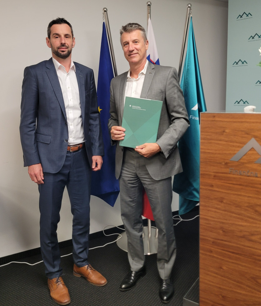  Pred zastavo EU, Slovenije in Finančne uprave stojita podpisnika sporazuma, Petkovšek v roki drži mapo v zeleni barvi finančne uprave