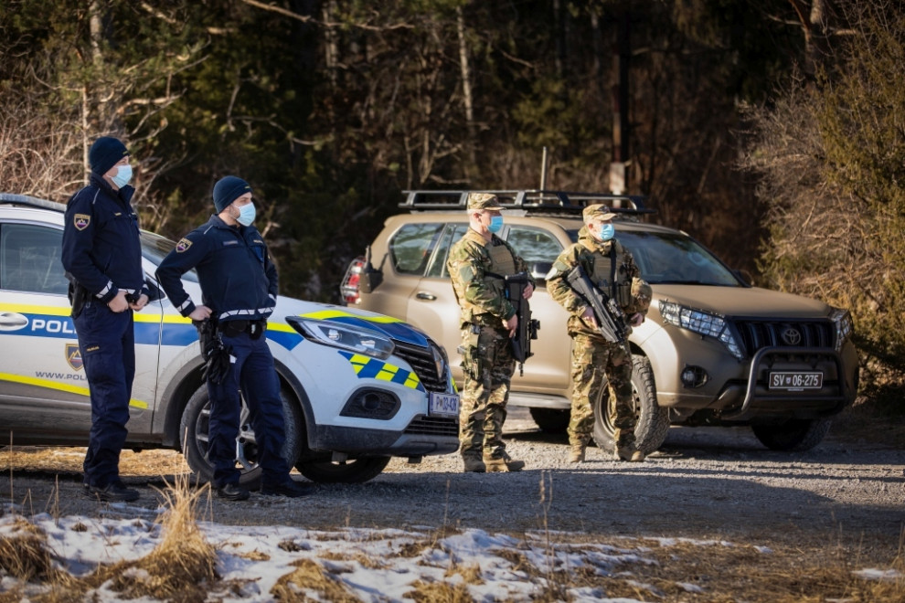 Dva policista in dva vojaka stojita pred policijskim in vojaškim vozilom pripravljena na nalogo patruljiranja ob meji.