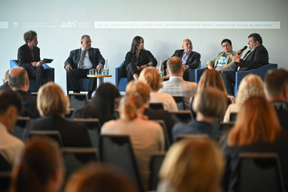 Skupina šestih predavateljev med panelno razpravo sedi na odru pred občinstvom med konferenco v Kongresnem centru Mons v Ljubljani, v ozadju je transparent, ki označuje organizatorja, naslov ter datum in lokacijo dogodka.