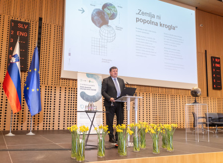 Govorec stoji za govorniškim pultom pred vazami rumenih rož, v ozadju slovenska in EU zastava in platno s prikazom predstavitve Zemlja ni popolna krogla