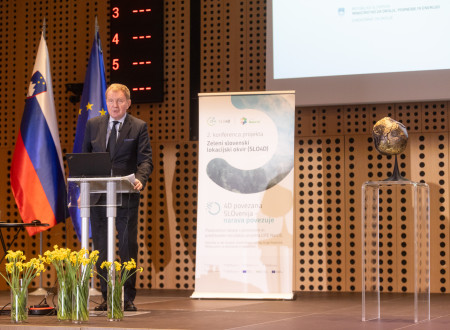 Govorec stoji pred govorniškim pultom, v ozadju zastavi Slovenije in EU ter pano projekta Zeleni slovenski lokacijski okvir