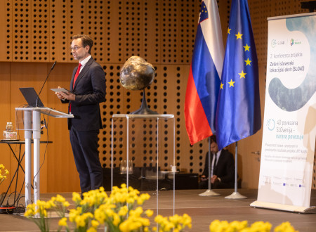 Govorec pred govorniškim pultom, pred njim maketa geoida Zemlje, v ozadju zastavi Slovenije in EU