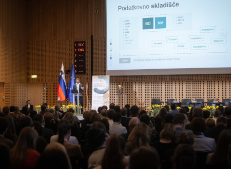 govorec stoji na odru, v ospredju dvorana s publiko, v ozadju platno z njegovo predstavitvijo