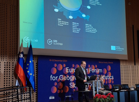 Govorec stoji za govorniškim pultom, v ozadju platno, kjer se prikazuje prezentacija njegovega prispevka
