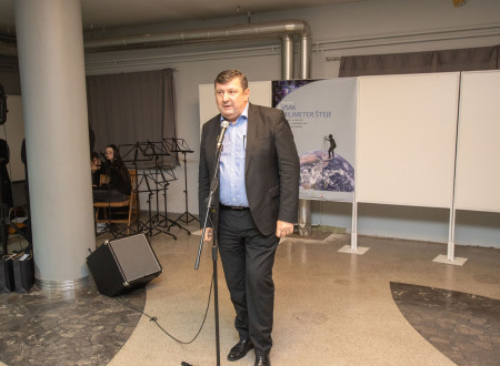Tomaž petek stoji pred mikrofonom, v ozadju razstavni panoji in učenci glasbene šole Trbovlje