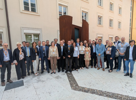 Na sliki stoji množica ljudi – udeležencev konference na trgu pred veliko stavbo z okni