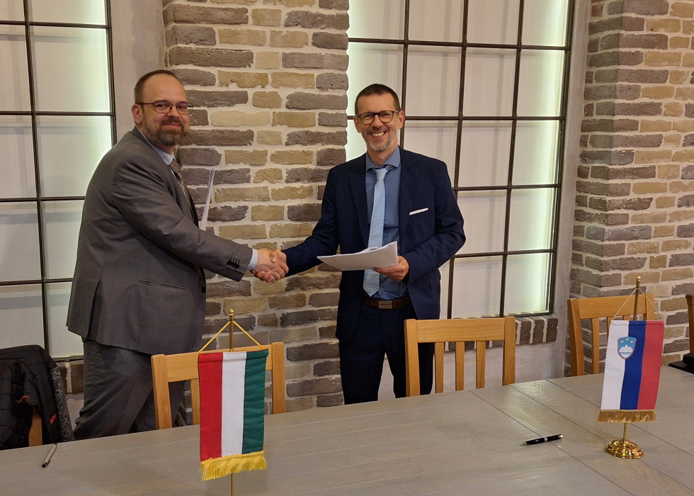 Dva moška, predstavnika Slovenije in Madžarske, se rokujeta ob podpisu zapisnika. Na mizi pred njima sta slovenska in madžarska namizna zastavica