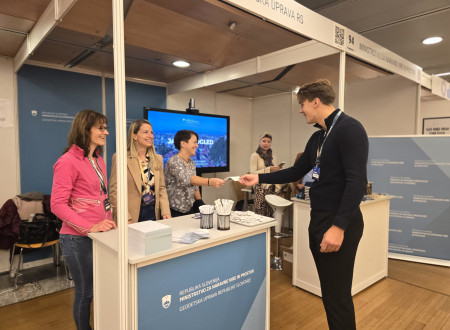 Tri predstavnice Geodetske uprave stojijo za pultom, ena izroča informativni letak mlajšemu obiskovalcu. Na pultu so publikacije in svinčniki.