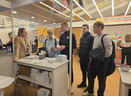 Na fotografiji predstavnica Geodetske uprave stoji za razstavno mizo in se nasmejano pogovarja s štirimi mladimi obiskovalci. Na mizi so zloženke in svinčniki, v ozadju so stojnice drugih razstavljavcev. Vsi so obrnjeni drug proti drugemu in držijo inform
