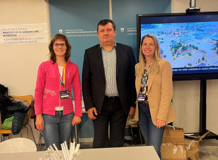 Pred razstavno mizo Geodetske uprave stojijo tri osebe, levo in desno predstavnici kadrovske službe Geodetske uprave, na sredini je direktor,  vsi gledajo proti fotoaparatu. Na mizi so zloženke in lončki s svinčniki; v ozadju je velik zaslon z grafiko, po