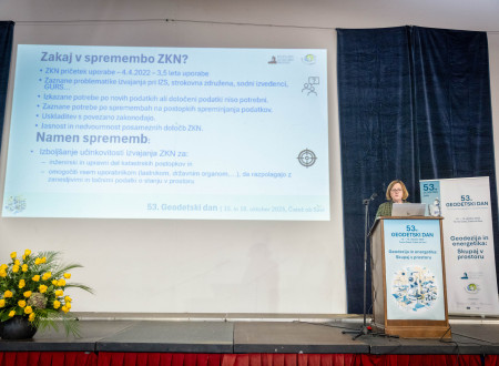 Predavateljica stoji ob pultu, na projekciji je naslov »Zakaj v spremembo ZKN?« z alinejami.