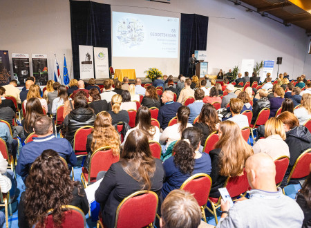 Širok posnetek dvorane z rdečimi stoli; na odru poteka predavanje s konferenčno projekcijo.