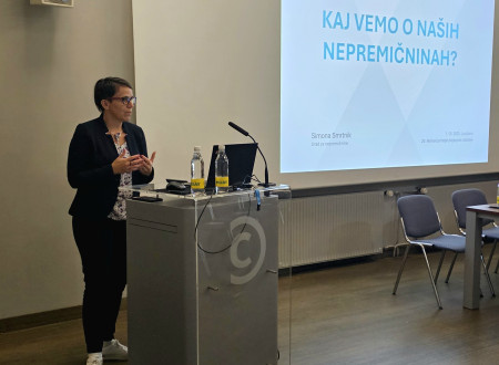 Simona Smrtnik stoji pred govorniškim odrom, v ozadju se na platnu prikazuje njena predstavitev.  