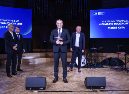 Moški v poslovni obleki stoji na sredini odra in v rokah drži nagrado, za njim stojijo trije moški ter glasbena oprema.