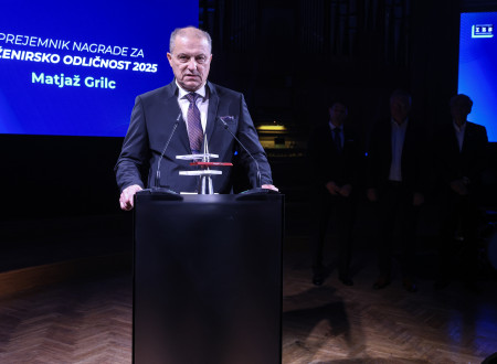 Moški v poslovni obleki stoji za govorniškim pultom z nagrado, v ozadju je projekcijsko platno z napisom o prejemniku nagrade.