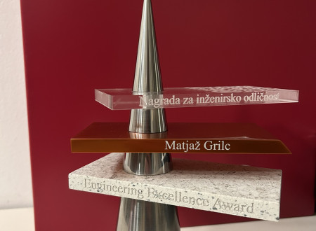 Kovinska in kamnita nagrada z vgraviranimi napisi stoji na beli površini pred enakomerno rdečim ozadjem.