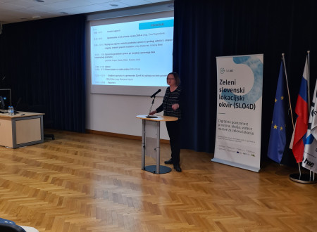 Velika skupina moških in žensk sedi v predavalnici in spremlja žensko predavateljico pri predstavitvi na projekcijskem platnu.