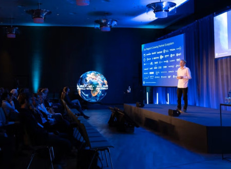 Moški stoji na odru pred veliko projekcijo in v dvorani nagovarja občinstvo med glavnim govorom.