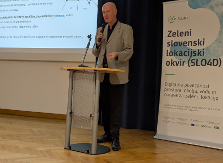 Moški predavatelj stoji za govorniškim pultom pred projekcijskim platnom s prikazom mej parcel, ob strani je pano projekta SLO4D. 