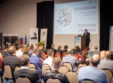 Govornik stoji za pultom pred polno dvorano; na projekciji piše »Slavnostna akademija«.