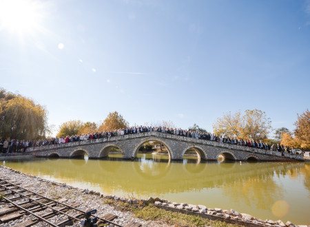 Velika skupina udeležencev stoji na kamnitem mostu nad jezerom v sončnem dopoldnevu; jesensko drevje in modro nebo v ozadju