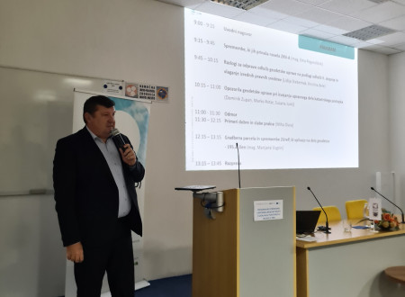 Moški predavatelj z mikrofonom stoji ob govorniškem pultu, na projekcijskem platnu za njim je prikazan program regionalnega strokovnega posveta.