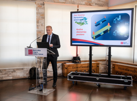 Moški v poslovni obleki predava za govorniškim pultom ob velikem zaslonu z diapozitivom o krožni ekonomiji prostora.