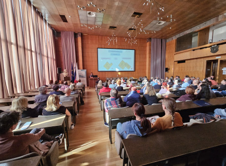 Skupina moških in žensk sedi v dvorani in posluša moškega predavatelja pred projekcijskim platnom in panojem SLO4D.