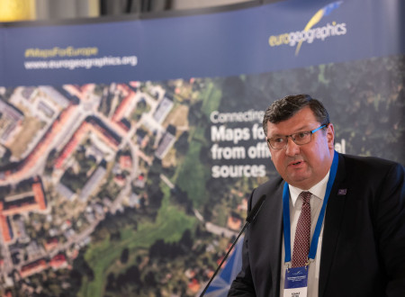 Moški v poslovni obleki govori na govorniškem odru pred panojem EuroGeographics.