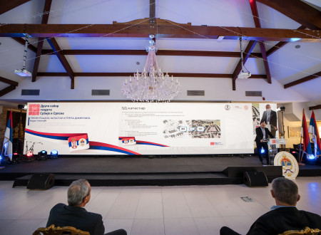 Širok pogled na oder z velikim projekcijskim zaslonom, na katerem je prikaz 3D katastra; moški v poslovni obleki stoji pri govorniškem pultu.