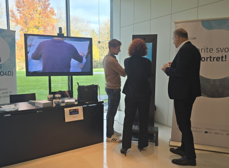 Obiskovalci preizkušajo interaktivni zaslon »Ustvari svoj portret«; na mizi promocijsko gradivo SLO4D, levo roll-up SLO4D, desno roll-up z napisom »Ustvarite svoj geoportret!«.